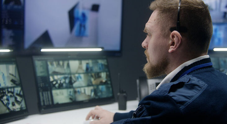 A security guard monitors multiple surveillance screens and operates a radio in the control room.