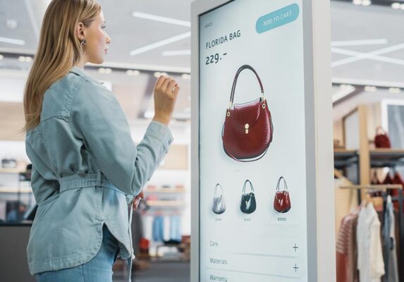 A person interacts with a touchscreen display in a store to view details of a red handbag.