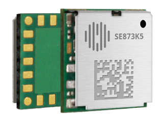 Close-up of a small electronic chip with a metal casing on one side and visible circuitry and connectors on the other side. The marking "SE873K5" is printed on the metal casing.