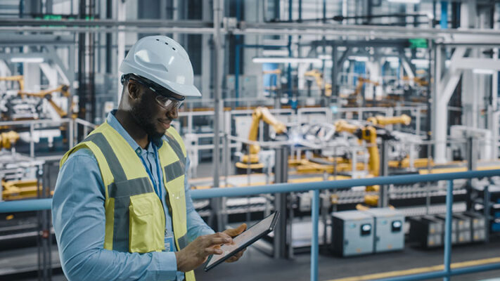 A worker in a hard hat and safety vest uses a tablet to oversee IIoT systems management in a modern factory with automated machinery in the background.