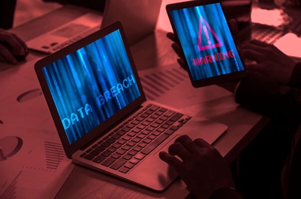 A person is using a laptop with the words "Data Breach" displayed and holding a tablet that shows a warning symbol.