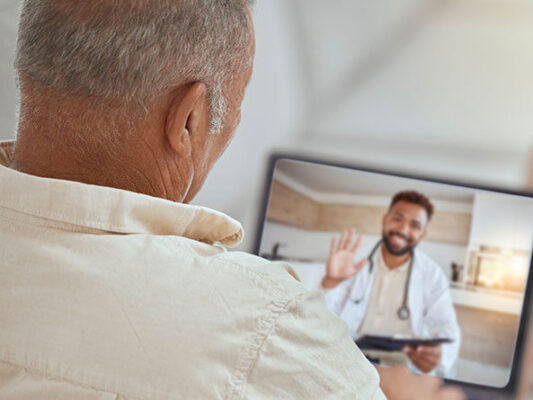 A person joins a connected health care video call with a doctor, who is smiling and waving. 