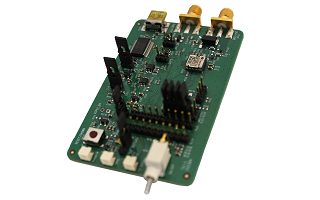 Close-up of a green circuit board featuring multiple components, including connectors, pins, chips, and a white button, with a small antenna-like protrusion at the bottom.