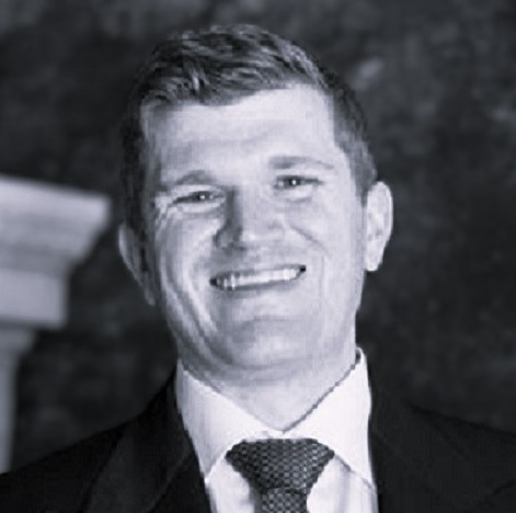 Smiling person in a suit and tie, posed against a dark, blurred background.