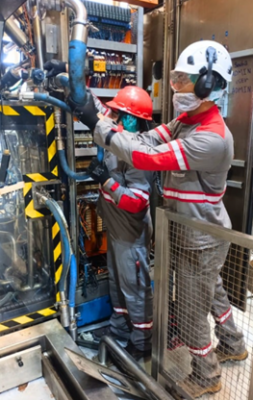 Two workers in protective gear, including helmets and masks, operate machinery with various cables and components in an industrial setting.