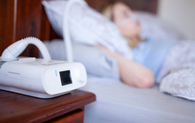 A person sleeps in bed while a CPAP machine, enhanced with edge AI for connected health care, sits on the bedside table.