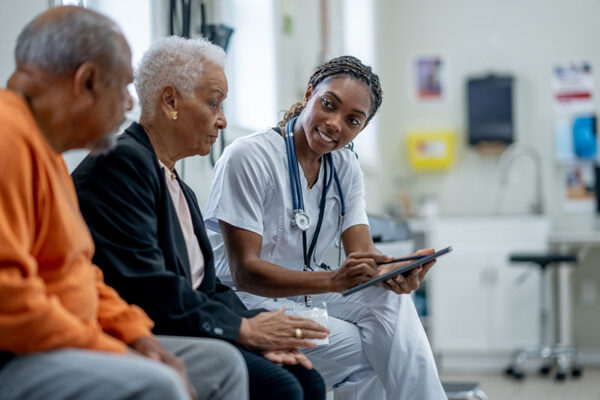 A healthcare professional in scrubs speaks with two older adults, holding a tablet and taking notes.