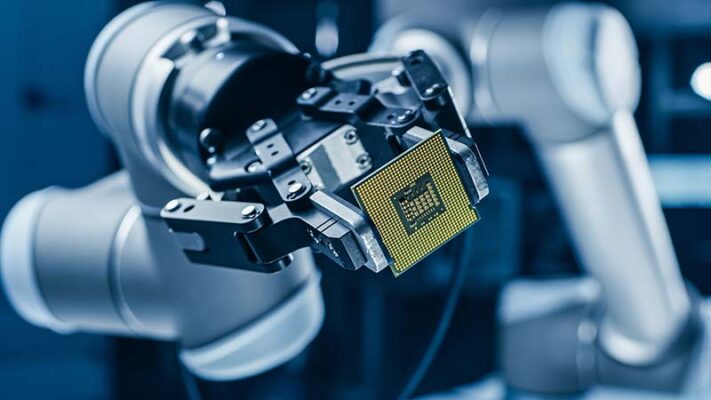 A robotic arm holds a gold computer processor chip between two clamps in a factory setting, where two-step personalization should occur.