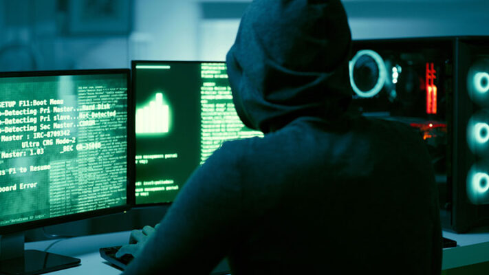 A person in a hoodie sits at a desk with multiple monitors displaying lines of code and data in a dimly lit room.