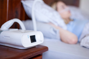 A CPAP machine sits on a bedside table, while a person sleeps peacefully in bed in the background.