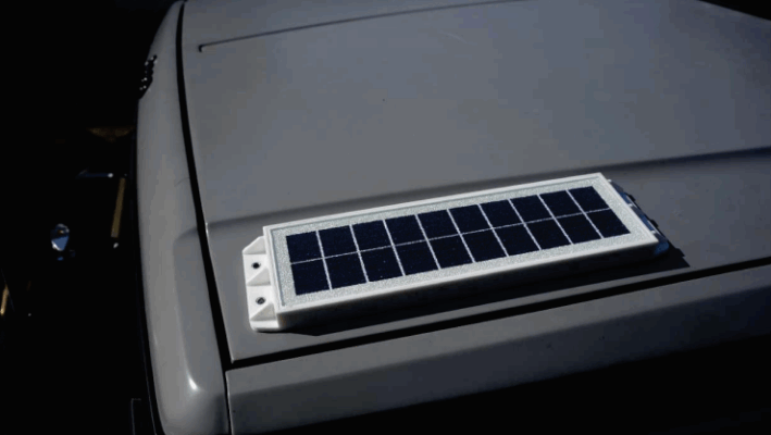 A small rectangular solar panel device with a telic design is mounted on the roof of a gray vehicle.