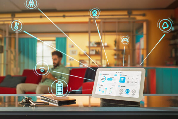 A smart home control panel sits on a table, displaying various settings and device icons, while a person relaxes on a couch in the background.