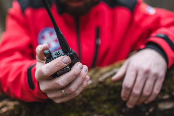 Person in a red jacket holds a walkie-talkie, resting their other hand on a mossy surface outdoors.