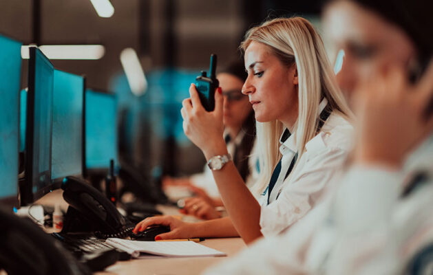 A person in uniform uses a walkie-talkie while working at a computer in a control room equipped with PERS technology. Other people are also seen at computer stations.