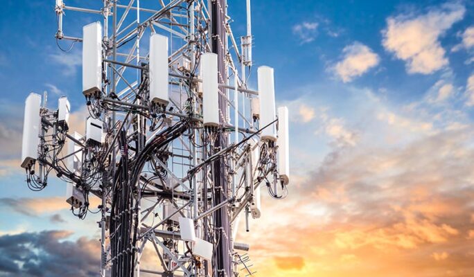 The top of a cellular tower with multiple antennas and cables set against a partly cloudy sky at sunset.