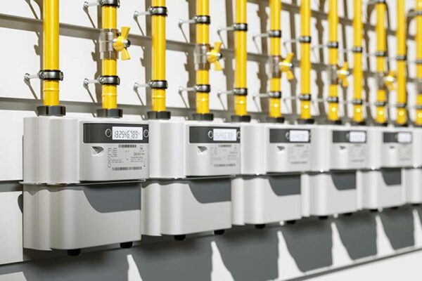 Five smart gas meters, each with two yellow pipes extending up, are mounted on a wall.
