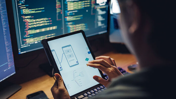 A person uses a tablet to design an IoT prototype with computer screens in the background displaying code.