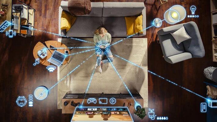 An overhead view of a person sitting in front of a living room couch holding a smart device and surrounded by digital icons.