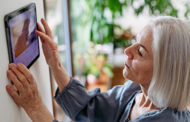 An older person with gray hair uses a wall-mounted touchscreen device.