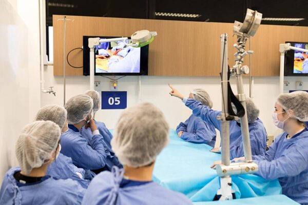 A group of medical students in surgical scrubs surrounds a hospital bed as they watch a procedure on a monitor.