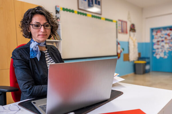 A woman works on a laptop at a desk in a classroom with a whiteboard and colorful decorations in the background.