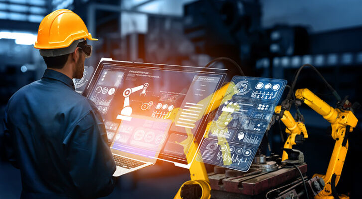 A worker in a hard hat operates a robotic arm using a laptop with digital displays in an industrial setting.