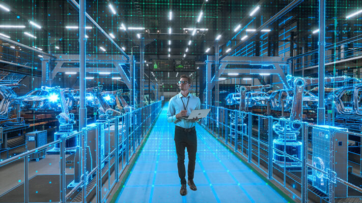A person holding a laptop walks through a modern factory with robotic arms, overlaid by blue digital and holographic interface graphics.