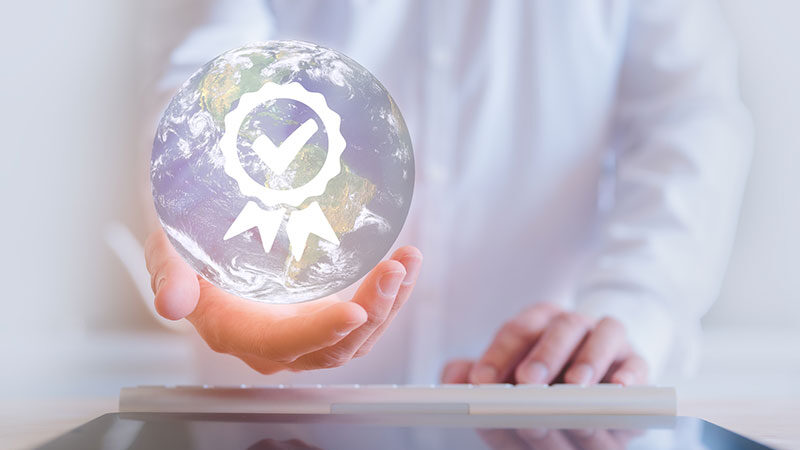 A person holds a transparent globe with a quality checkmark symbol while typing on a keyboard, representing the search for the right IoT module provider.