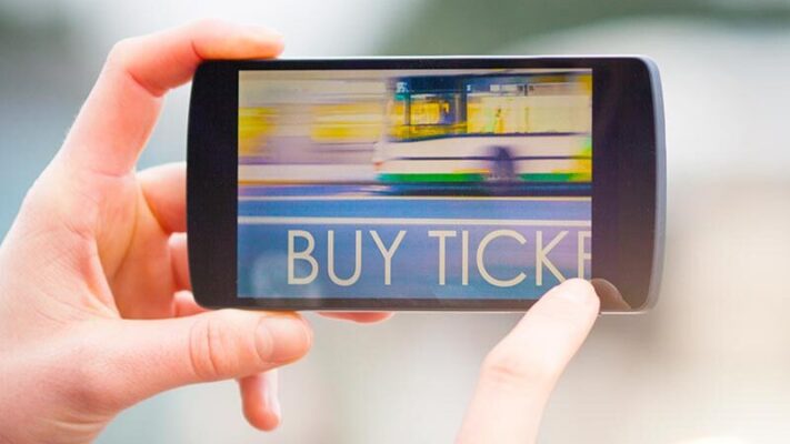 A person holds a smartphone displaying a blurred image of a bus with the text "BUY TICKETS" on the screen.