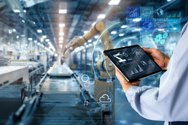 A person holds a tablet displaying data analytics in an automated smart factory with robotic arms on an assembly line.
