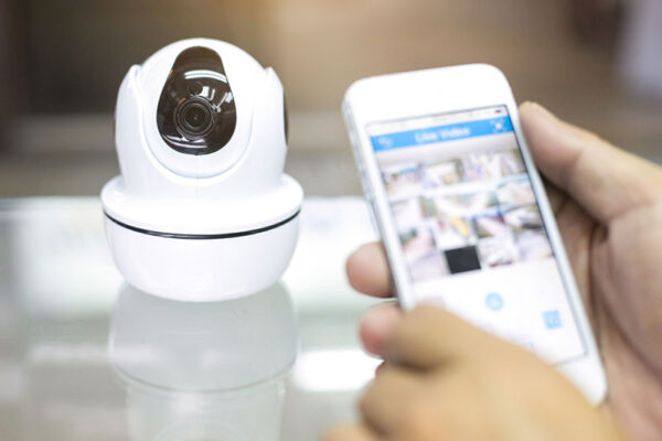 A person holds a smartphone displaying video surveillance footage next to a white security camera, which sits on a glass surface.