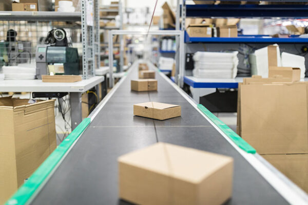 Small cardboard boxes move along a conveyor belt in a warehouse, representing a supply chain.