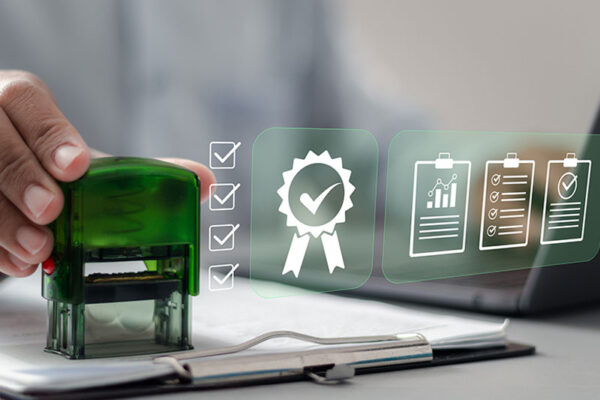 A person presses a green stamp onto a document next to digital icons of checklists and a certificate, symbolizing approval.