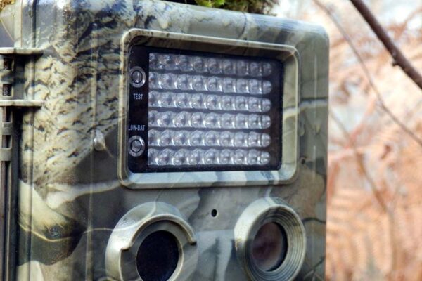 Close-up of a camouflaged smart trail camera with infrared LED lights, a test button and a low battery indicator mounted outdoors.