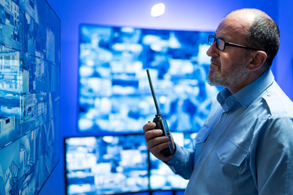A man holds a walkie-talkie while monitoring multiple security and surveillance screens in a control room.