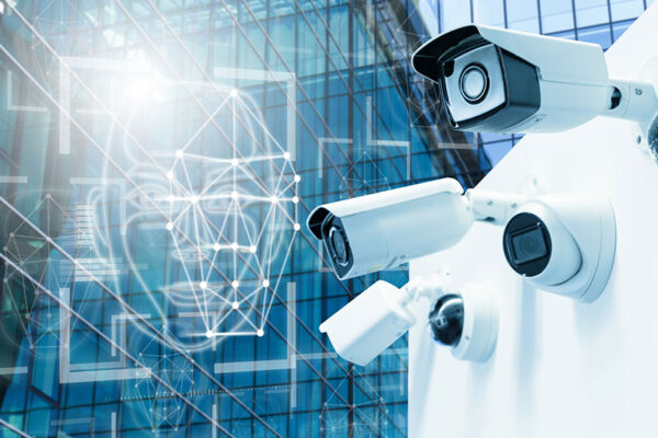 A digital rendition of facial recognition overlays a glass building next to a row of security and surveillance cameras mounted on an outdoor wall.