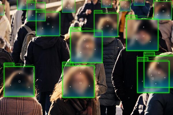 A crowd of people walking outdoors, with their faces blurred and outlined by green boxes, indicating the use of surveillance technology and facial recognition.