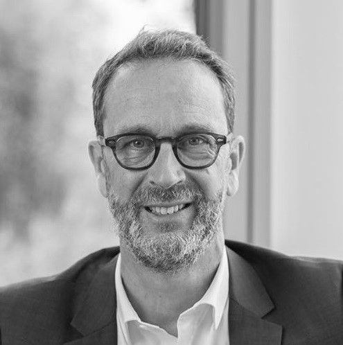 A middle-aged man with glasses, a beard, and short hair is smiling while wearing a suit jacket and shirt, seated in front of a window.