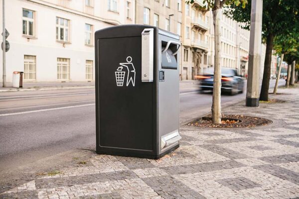 A smart city trash bin stands on a patterned sidewalk beside a street with blurred passing cars and buildings.