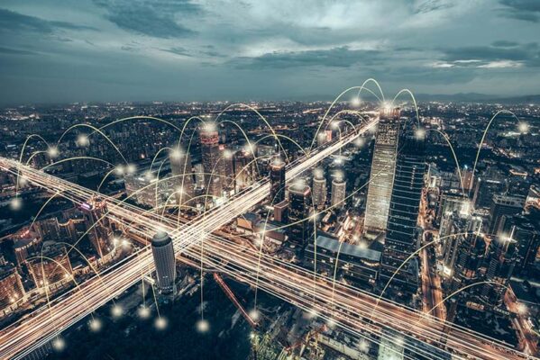 Aerial view of a city at dusk with light trails and digital lines representing data connections between skyscrapers and roads.