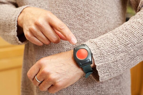 A person wearing a beige sweater points to a black wristband with a large red emergency button.