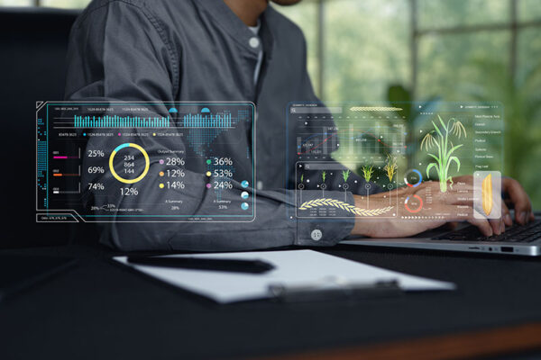 A person works on a laptop. Clear digital screens show farm data analytics. A notepad and pen sit on the desk.
