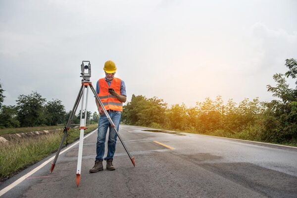 A worker wearing a yellow hard hat and an orange vest uses survey equipment on a tripod on an empty road, with trees and a cloudy sky in the background.