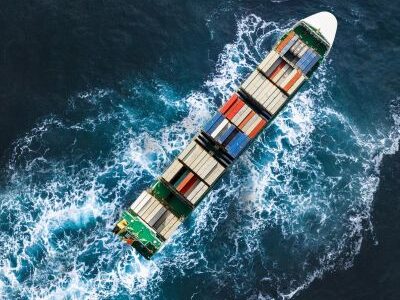 A cargo ship carrying containers moves through the ocean, leaving a wake behind it.