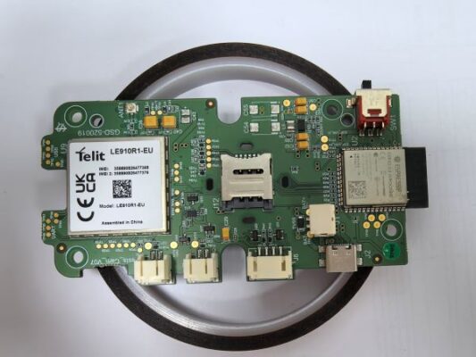 A green circuit board with multiple microchips, ports, and a Telit LE910R1-EU module, placed on a white background with a black ring underneath.