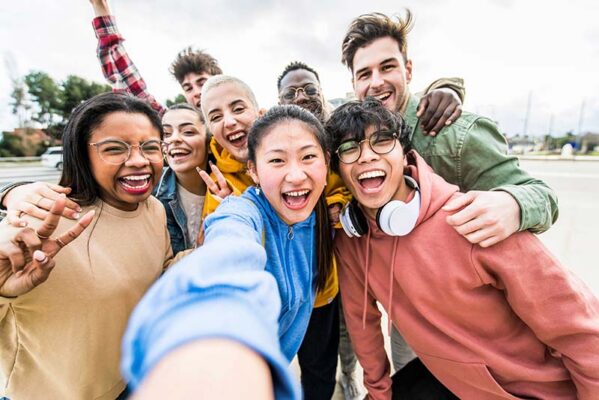 A group of eight diverse young people laughing and smiling while taking a selfie together outdoors.