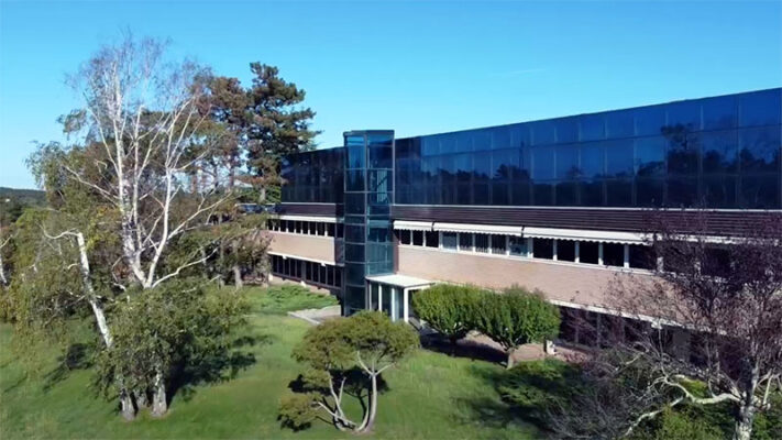 A two-story commercial building with a glass curtain wall, brick lower level, and a central glass elevator shaft, surrounded by trees and lawn.